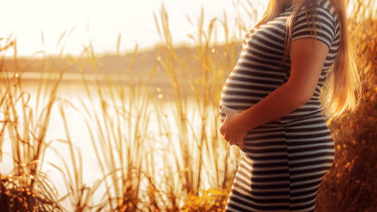 怀孕的妇女在日落时抚摸她的腹部. 一条河边的条纹连衣裙上的孕妇特写