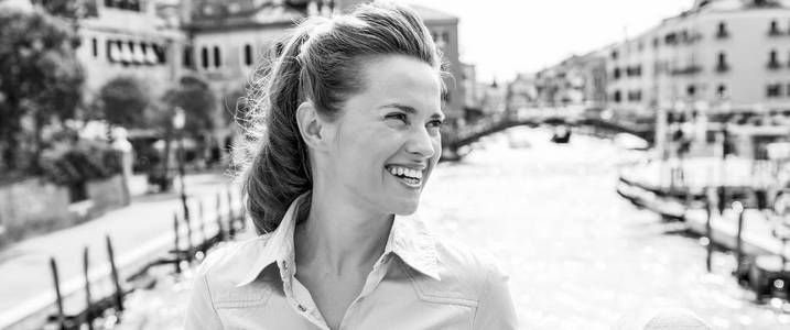 一个漂亮的黑发游客斜倚在威尼斯运河上的一座桥上, 微笑着望着远方。她是放松和愉快的