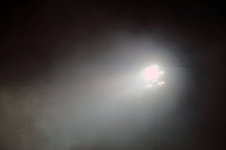 黑夜期间, 一座体育场点燃了灯光塔。体育场灯反对黑暗的夜空背景。体育场灯和烟雾