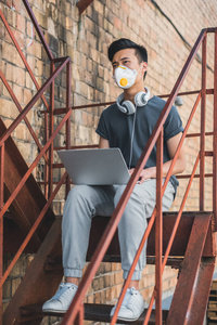 亚洲青少年在防护面具坐与膝上型电脑在楼梯和看, 空气污染概念