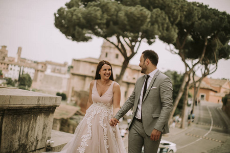 年轻迷人新婚夫妇步行和摆在罗马与美丽的古建筑在他们的婚礼当天的背景
