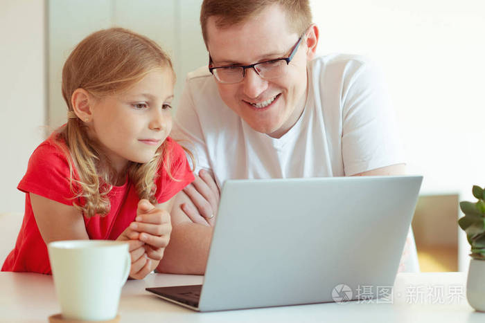 愉快的年轻的父亲有乐趣与他漂亮的小女儿在工作与笔记本电脑在现代