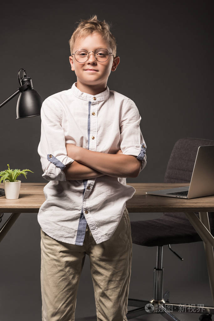 小男孩在眼镜与交叉的手站立在桌附近与植物,膝上型电脑和灯在灰色