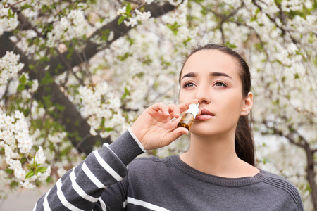 年轻女子用鼻滴在盛开的树附近。过敏概念