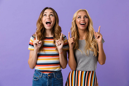 两个漂亮的年轻女孩的朋友站在紫色背景隔绝, 指着手指向上