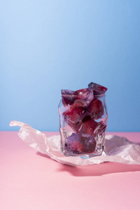 冻红浆果在玻璃上的皱巴巴的包装纸站在粉红色的桌子上的蓝色背景。概念特写照片。复制空间。广告与商业设计