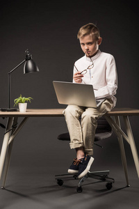 严肃的小男孩拿着眼镜和使用膝上型电脑, 而坐在桌子上与植物和灯的灰色背景