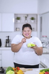 在厨房里吃一碗健康的沙拉时, 胖女人的照片看起来很快乐。