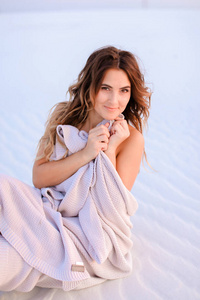 美丽的女性人裹着毯子坐在白色的沙子上