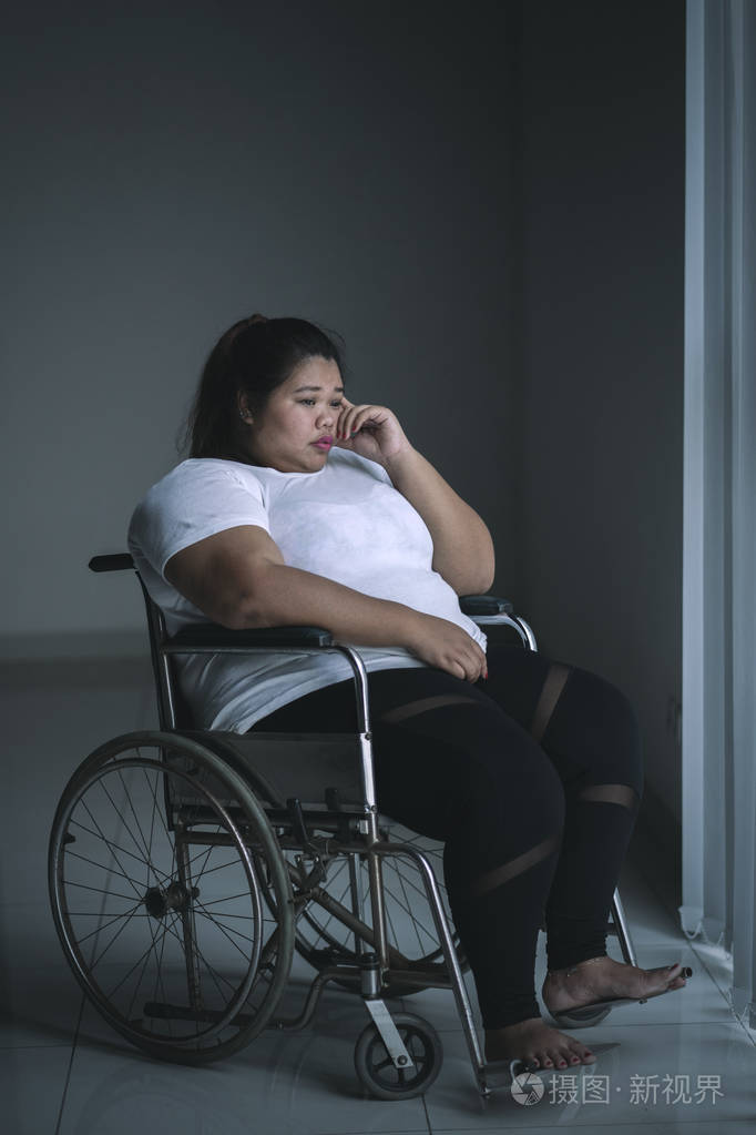 残疾肥胖妇女纵向坐在轮椅旁边的窗口看起来很悲伤