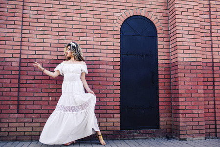 美丽的女孩穿着白色礼服站在墙边的金属门和摆姿势
