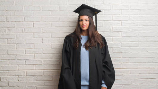 年轻的黑发女子站在白色砖墙穿着毕业生制服与自信的表情在聪明的脸上思考严肃