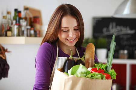 年轻女子捧着与蔬菜站在厨房里的食品杂货店购物袋