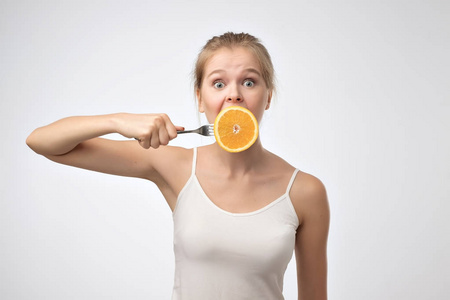 美丽的女人在白色的衬衫拿着一个叉子与半橙色靠近她的嘴, 而站立反对白色背景