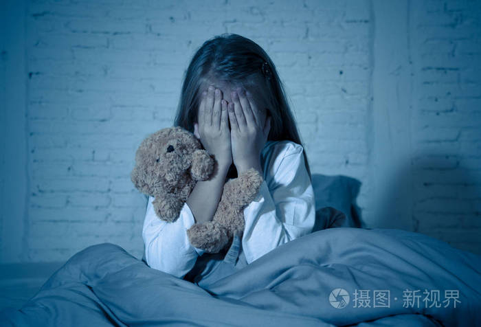 害怕的小女孩坐在床上用双手抱着她的泰迪熊捂着她