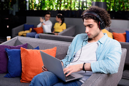 年轻的商人在耳机坐在沙发上与笔记本电脑在现代 coworking 办公室