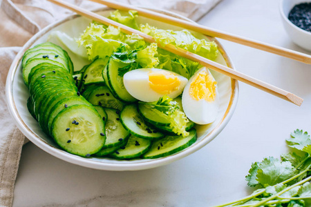 大理石背景下配有筷子的黄瓜沙拉和鸡蛋碗照片