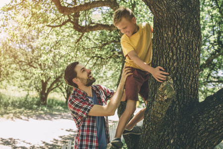 有胡子的家伙站在树边支持他的儿子。男孩正在爬起来。孩子把手放在树上, 看着他的父亲。他们在微笑