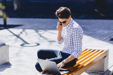 年轻的时尚男子与电话工作在笔记本电脑在公园阳光明媚的一天