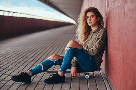 穿着帽衫和撕裂牛仔裤倚在墙上, 而坐在一个滑板在桥人行道照片
