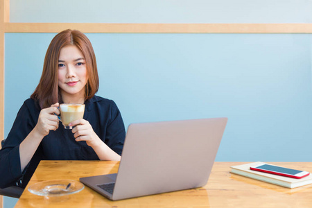 快乐的年轻的亚洲商人享受她的热咖啡, 而在她的办公室工作, 在她的电脑, 良好的现代工作生活方式或咖啡的概念
