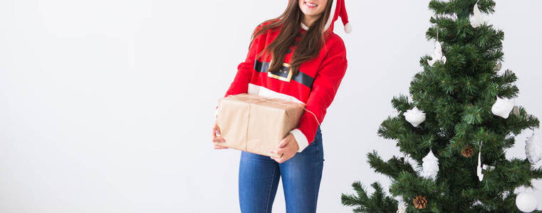 年轻开朗的女人拿着礼品盒穿着圣诞老人的服装。圣诞树背景与拷贝空间