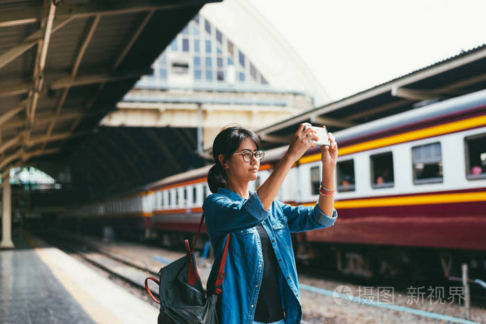 亚洲女旅行者在火车站站台上等待和查看火车时刻表时使用智能手机拍照