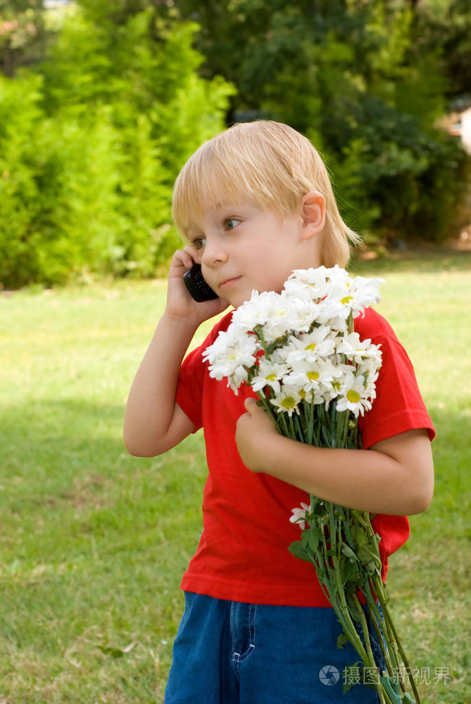 快乐的小男孩捧着白花, 电话站在绿草的背景上说话
