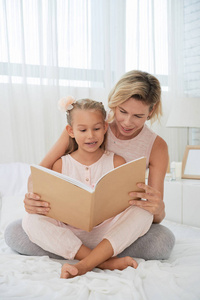 愉快的母亲和女儿一起读一本书在床在卧室
