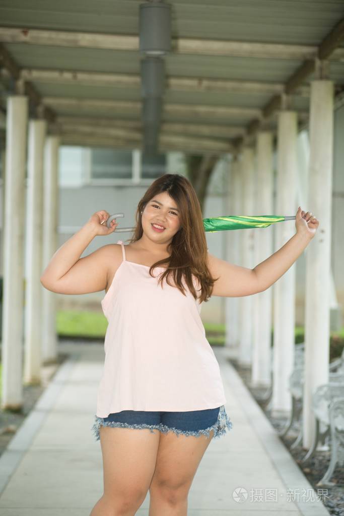 亚洲漂亮的笑脸脸胖女人姿势站立, 并持有雨伞在人行道上