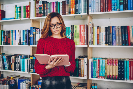 聪明的女记者搜索信息站在文学与书架上背景, 有魅力的年轻女子眼镜阅读畅销书在大学大学