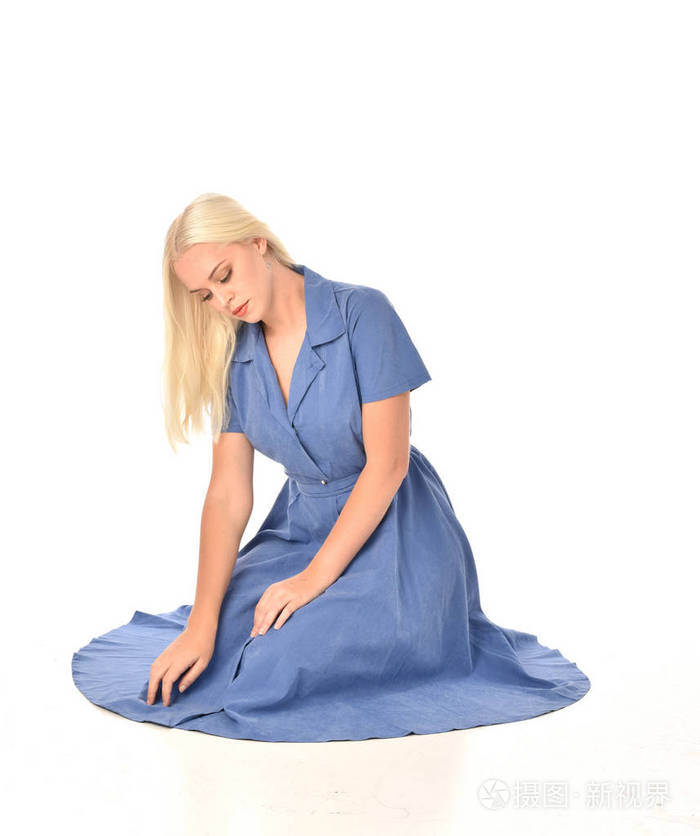 全长肖像的金发女郎穿着蓝色的礼服坐姿在白色工作室背景下被隔离