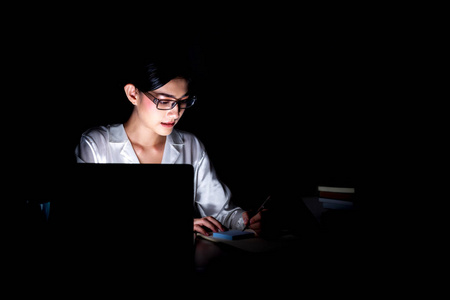 在她家办公室工作到很晚的女人。美丽的亚洲妇女工作在她的电脑, 写笔记, 戴眼镜。现代全球互联业务理念