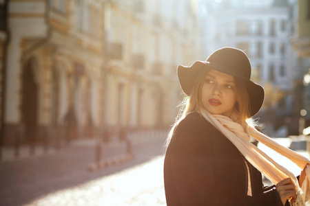 时尚的金发女子戴着帽子, 围巾和大衣, 走在老街上的太阳光线。文本空间