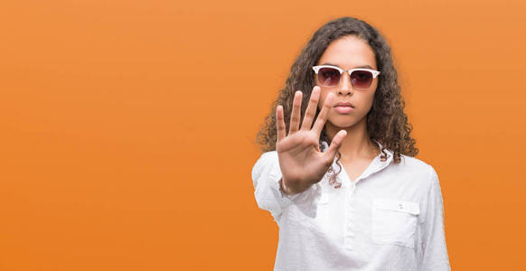年轻的西班牙裔妇女戴着墨镜, 张开手做停止标志与认真和自信的表达, 防御姿态