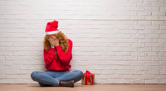 年轻的红头发的女人坐在砖墙上戴着圣诞帽, 哭的时候用手悲伤地表情着脸。抑郁症的概念