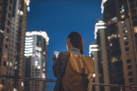 背面的妇女与背包和智能手机与购物标志在屏幕上的手站在夜间城市街