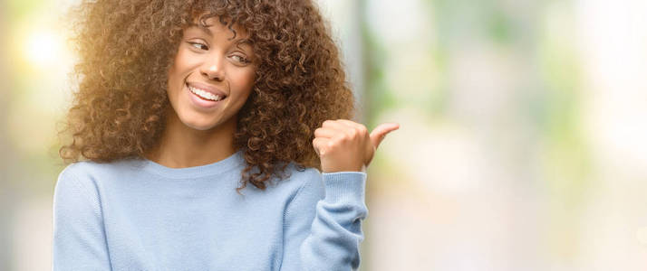 非洲裔美国妇女穿着一件毛衣微笑着快乐的脸, 用拇指指向侧面