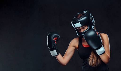 运动的女人穿着运动服, 戴着防护头盔和拳击手套, 在健身房训练。在深色纹理背景下隔离