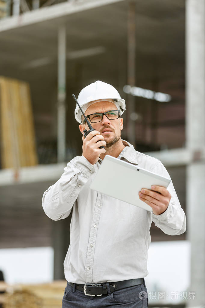 男总工程师戴着白色安全安全帽, 配有对讲机和平板电脑检查施工现场.