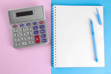 计算器和笔记本上的钢笔在蓝色和粉红色的背景。从上面查看。文具