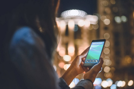 妇女的部分看法使用智能手机与预订徽标在屏幕上的夜晚城市灯光背景