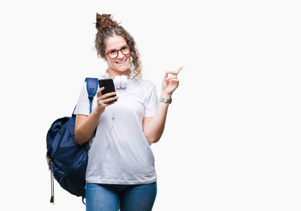 年轻的黑发学生女孩穿着背包, 耳机和智能手机在孤立的背景非常高兴地指着手和手指的一侧