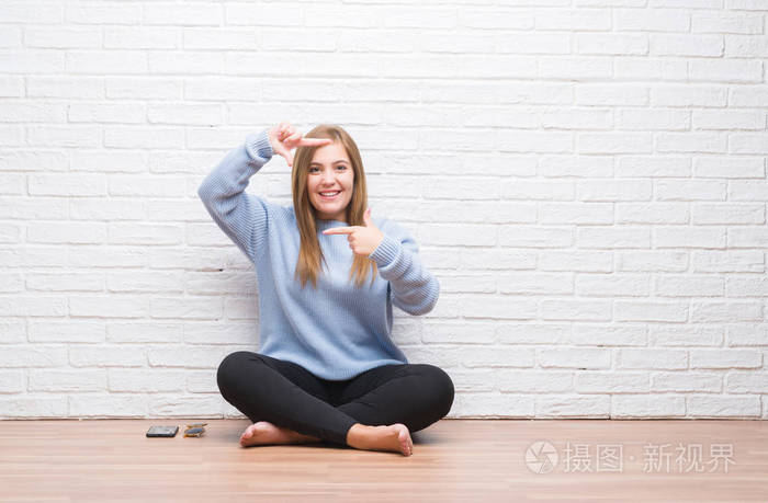 一位年轻的成年女子坐在地板上, 在秋天的白色砖墙上, 微笑着用手和手指与快乐的脸做框架。创意与摄影理念