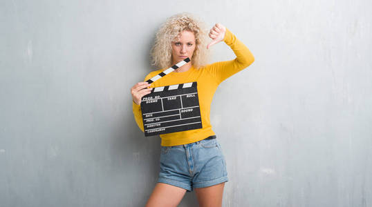 年轻的金发女人在垃圾灰色墙壁持有 calpboard 的电影与愤怒的脸, 否定的标志显示不喜欢拇指下来, 拒绝概念