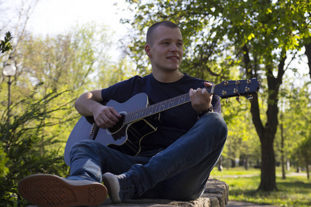 英俊的年轻人在公园里玩耍, 坐着弹吉他。选择性对焦和小景深