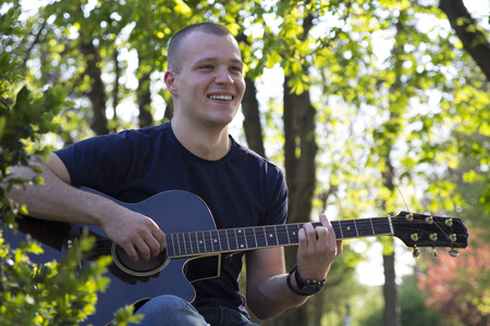 微笑的英俊年轻人喜欢在公园里弹吉他。选择性对焦和小景深