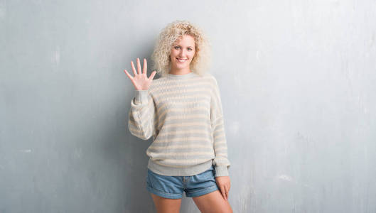 年轻的金发女人与卷曲的头发在垃圾灰色背景显示, 并指出手指五, 而微笑着自信和快乐