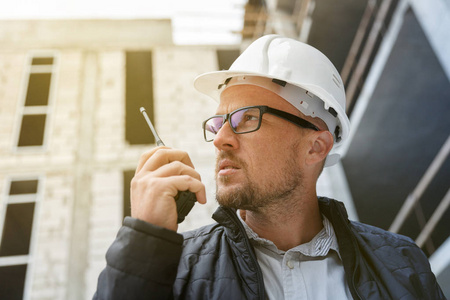 男性总工程师在检查施工现场时, 使用对讲机佩戴白色安全安全帽。发展和建筑业理念