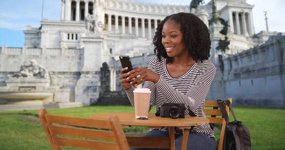 微笑的黑人妇女坐在威尼斯广场外的桌子上, 用智能手机发短信